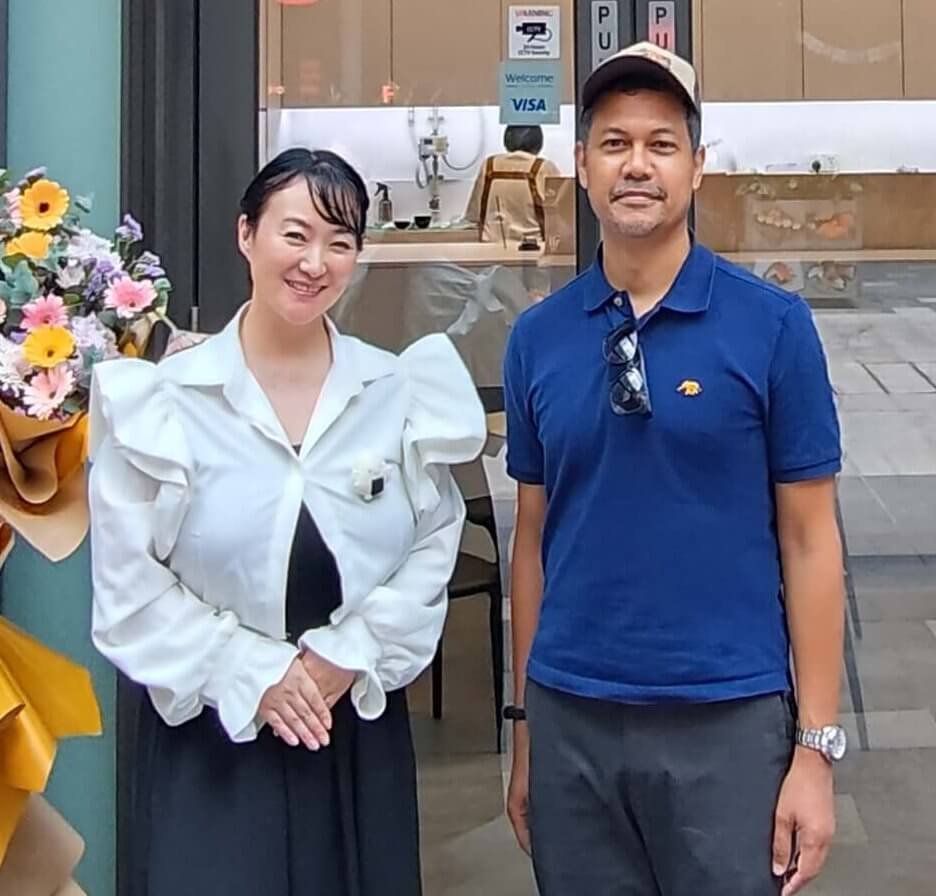 カフェの前で花束と共に立つ深瀬マリー麻美とイクバル・アバドゥラ。左の女性は白いフリルブラウスに黒いスカート、右の男性は青いポロシャツにサングラスを首にかけている。