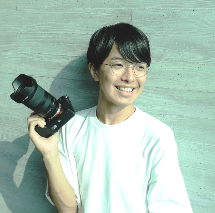 小澤 健祐 さんがカメラを手に笑顔を見せるポートレート。白いシャツを着ており、背景にはテクスチャのあるグレーの壁が見えます。