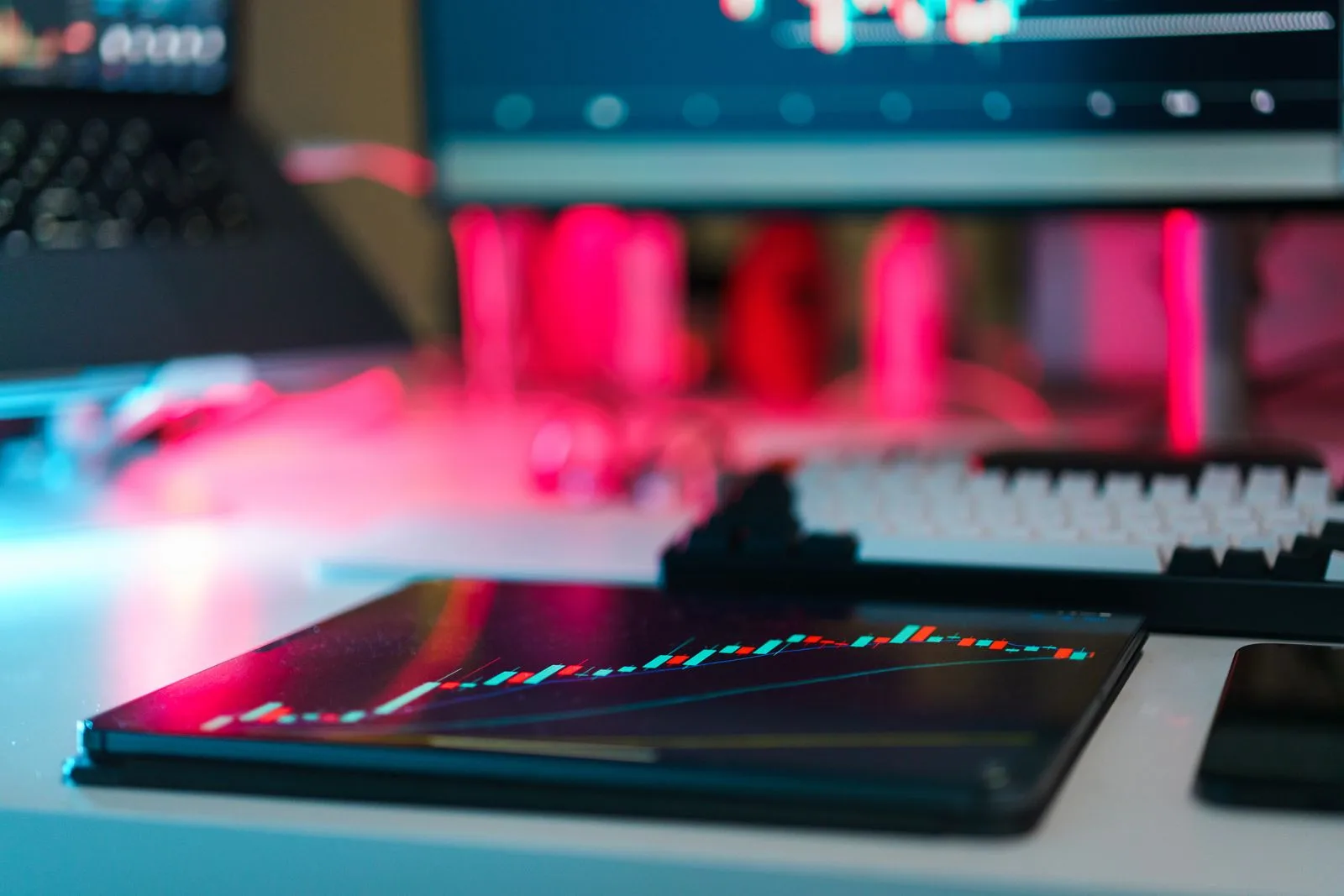 Tablet displaying a financial stock market graph with red and green candlesticks on a desk with blurred monitors.
