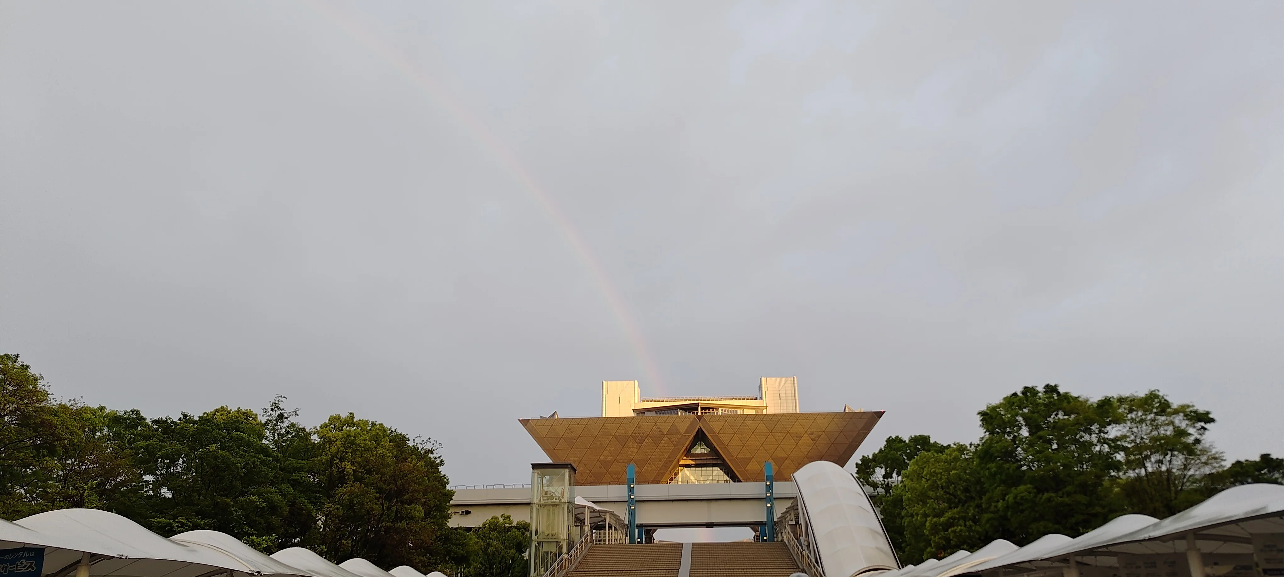 A photograph of a modern architectural building with a gold peaked roof under a cloudy sky with a faint rainbow.